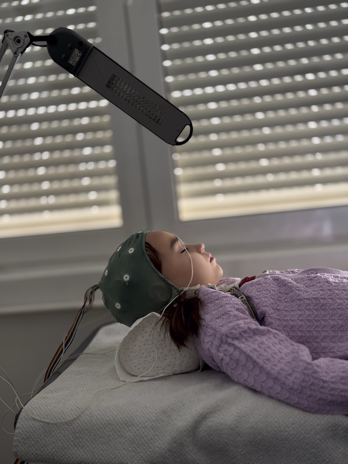 Criança deitada em maca de clínica com eletrodos de EEG na cabeça; a sala é iluminada e a criança parece relaxada durante o exame.