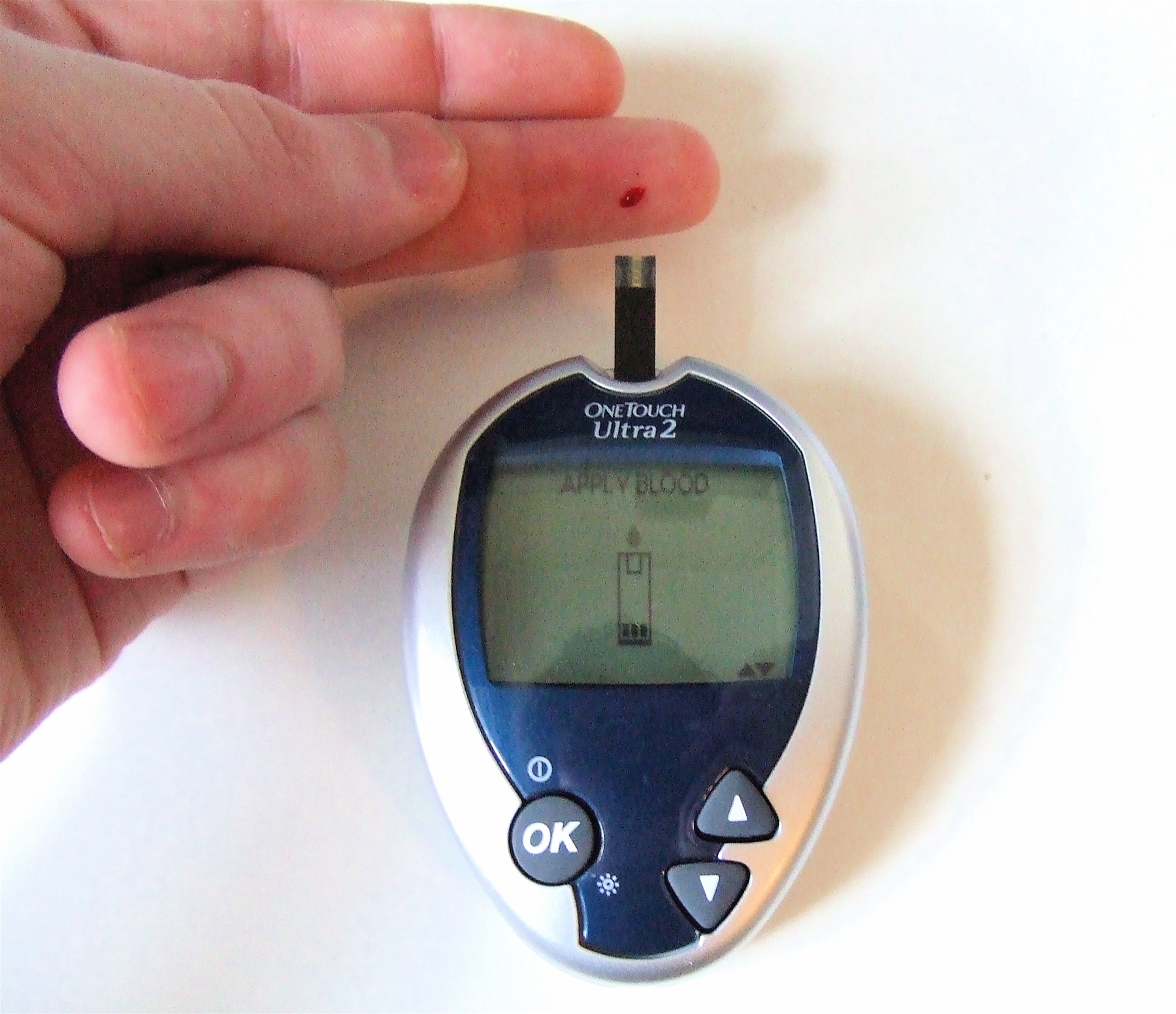 Glicosímetro portátil OneTouch Ultra 2 com tira reagente inserida e gota de sangue capilar no dedo, pronto para medição de glicemia; tela do aparelho mostra a instrução APPLY BLOOD.