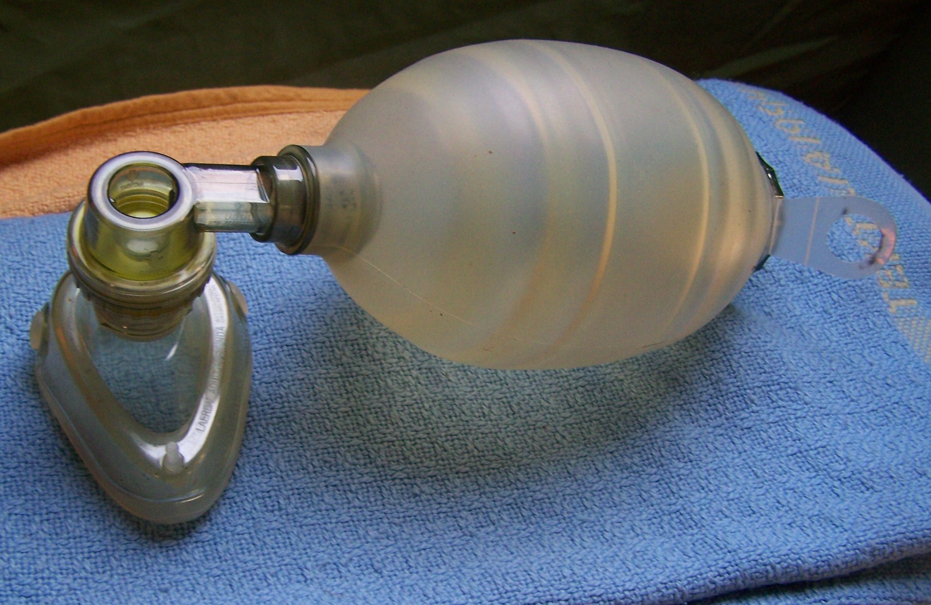 Bolsa-válvula-máscara (AMBU) azul e transparente sobre fundo branco, usada para ventilação manual de emergência.