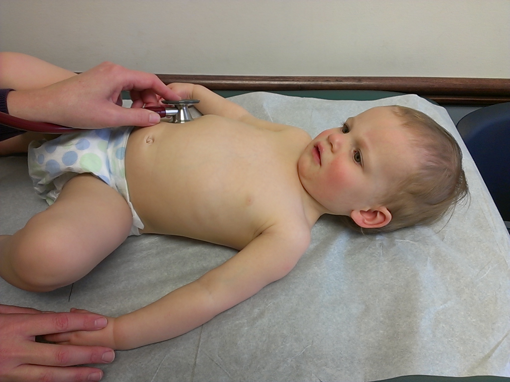 Criança pequena deitada em maca de consultório, com a mão de um profissional usando estetoscópio sobre o tórax.