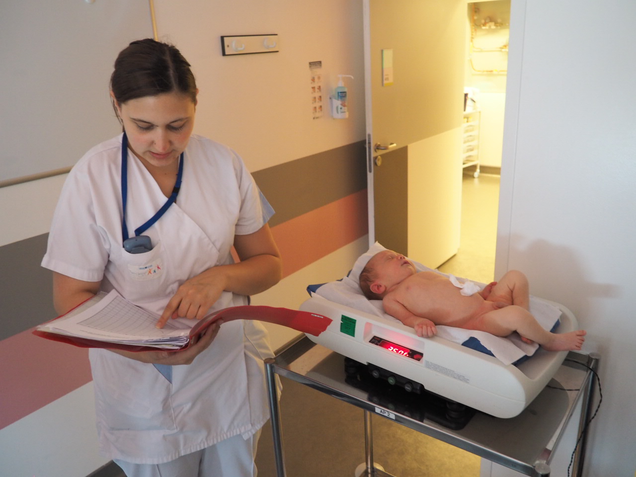 Enfermeira anotando dados em uma ficha enquanto um bebê é pesado deitado em uma balança pediátrica eletrônica de mesa, em hospital.