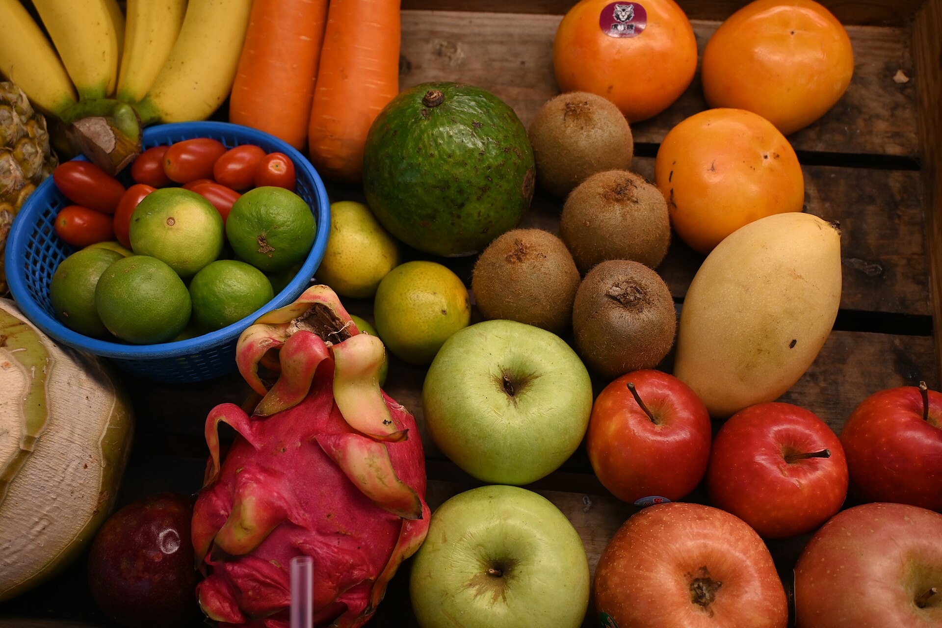 Composição de frutas e hortaliças frescas — cenoura, kiwi, limão, tomate, caqui, laranja, abacate, maçã, banana, manga e pitaia — sobre engradado de madeira, ilustrando alimentos-fonte das vitaminas.