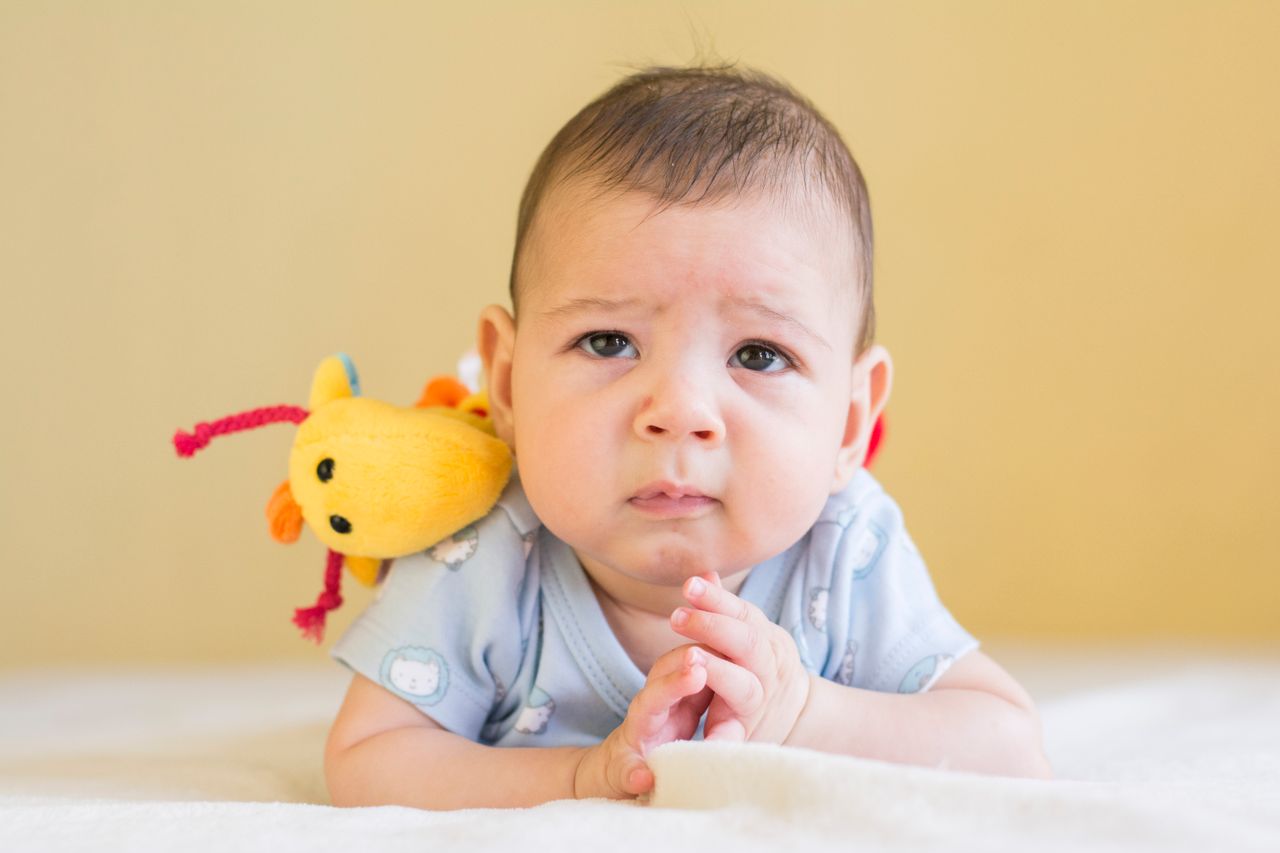 Foto real de um bebê saudável de bruços (prono), em tummy time, com um brinquedo macio ao lado.